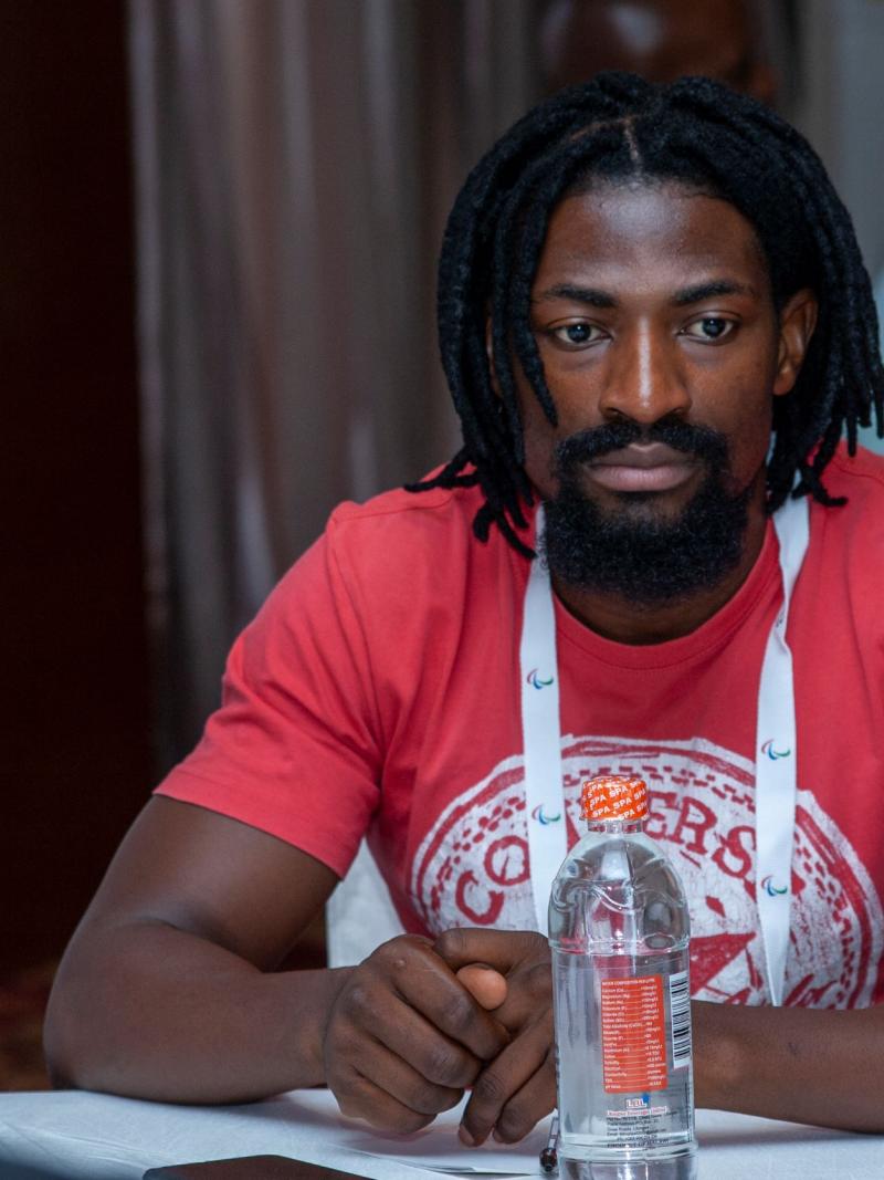 A man in a red t-shirt looks on as he sits at a table with his hands folded in front of him.