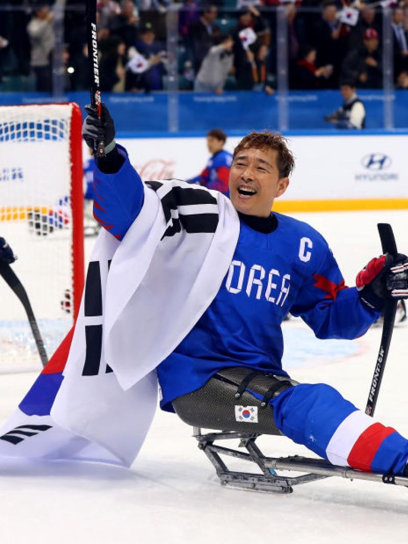 A male Para ice hockey player holds up a sledge hockey stick with his right hand and carries the Republic of Korea flag.
