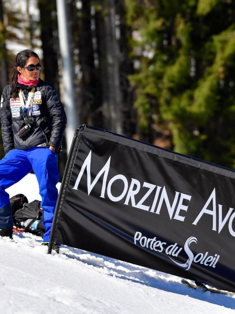 A female coach stands on the snow, next to a male staff and a sign that reads "Morzine Avoriaz"