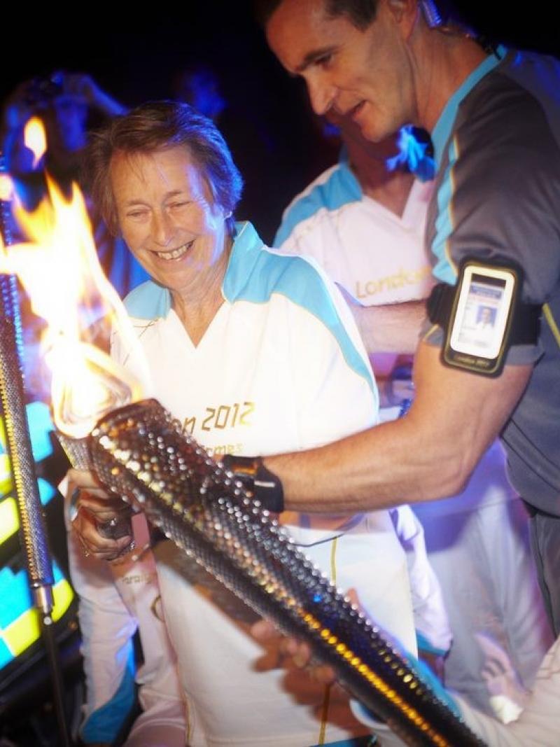 A woman in a wheelchair passes the Paralympic Flame through a torch to another woman, also holding a torch.