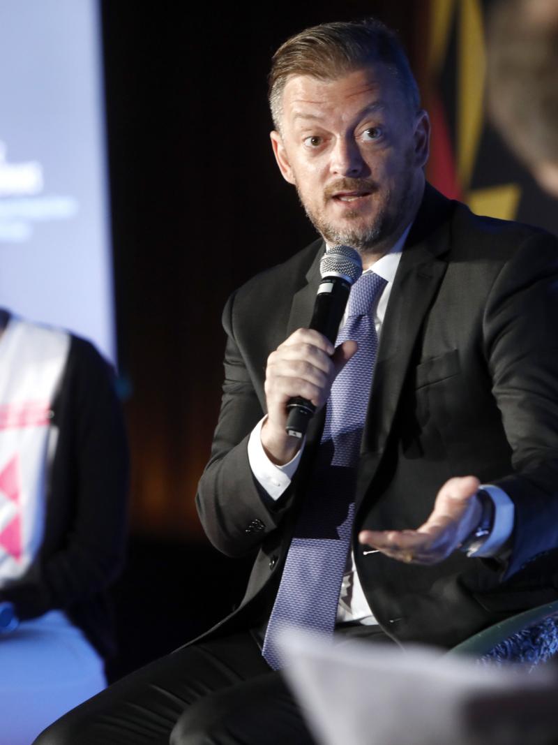 A man in a suit speaks into a microphone during a discussion session at Global Sport Week in Paris.