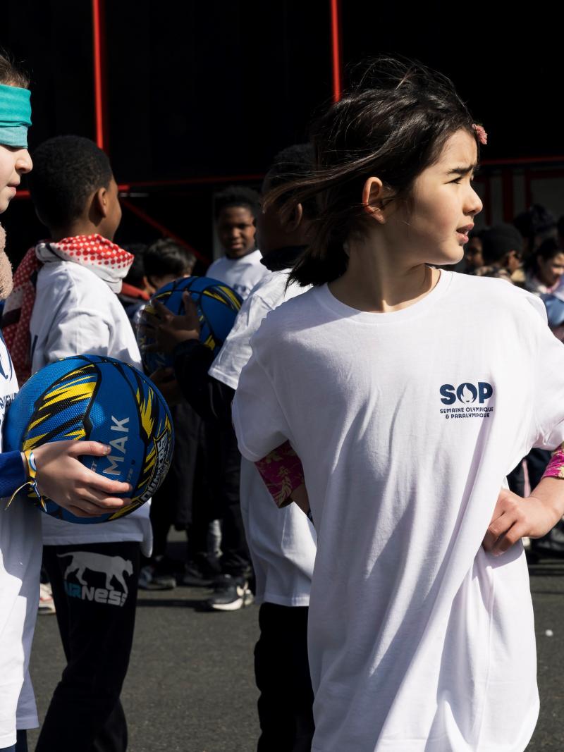 Two young girls, one with a blindfold, holding a ball, and the other looking back, participate in an event during Olympic and Paralympic Week.