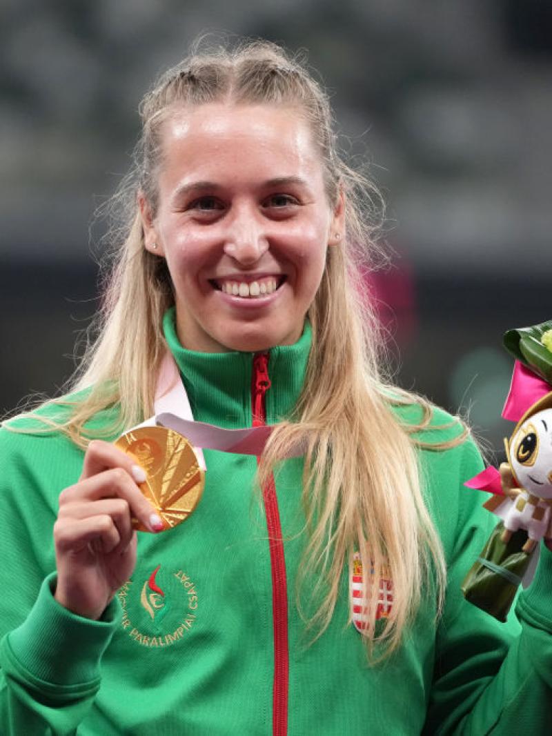 A female athlete smiles for a photograph while holding a gold medal with her right hand and a bouquet with her left hand.