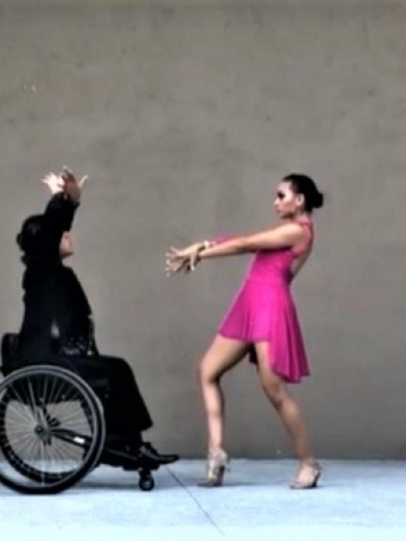A male wheelchair dancer dressed in black dancing together with a standing woman in a pink dress