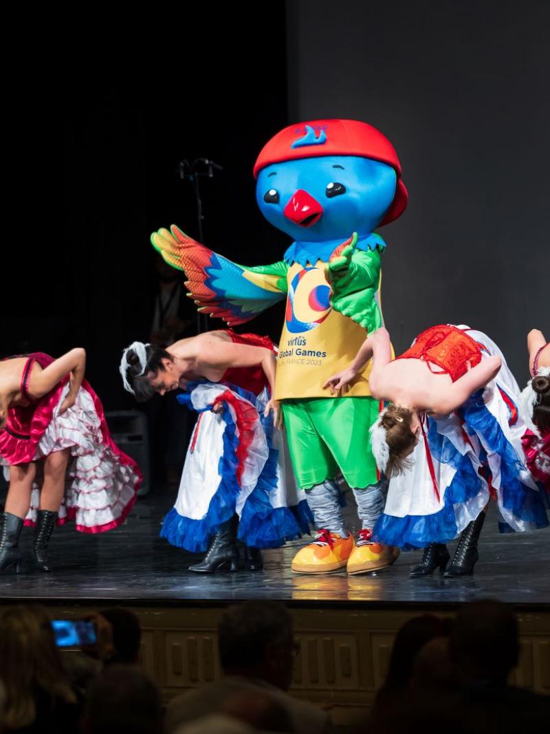 The mascot of the Virtus Global Games, which is a blue bird wearing a red cap, on a stage with seven women who are bowing.