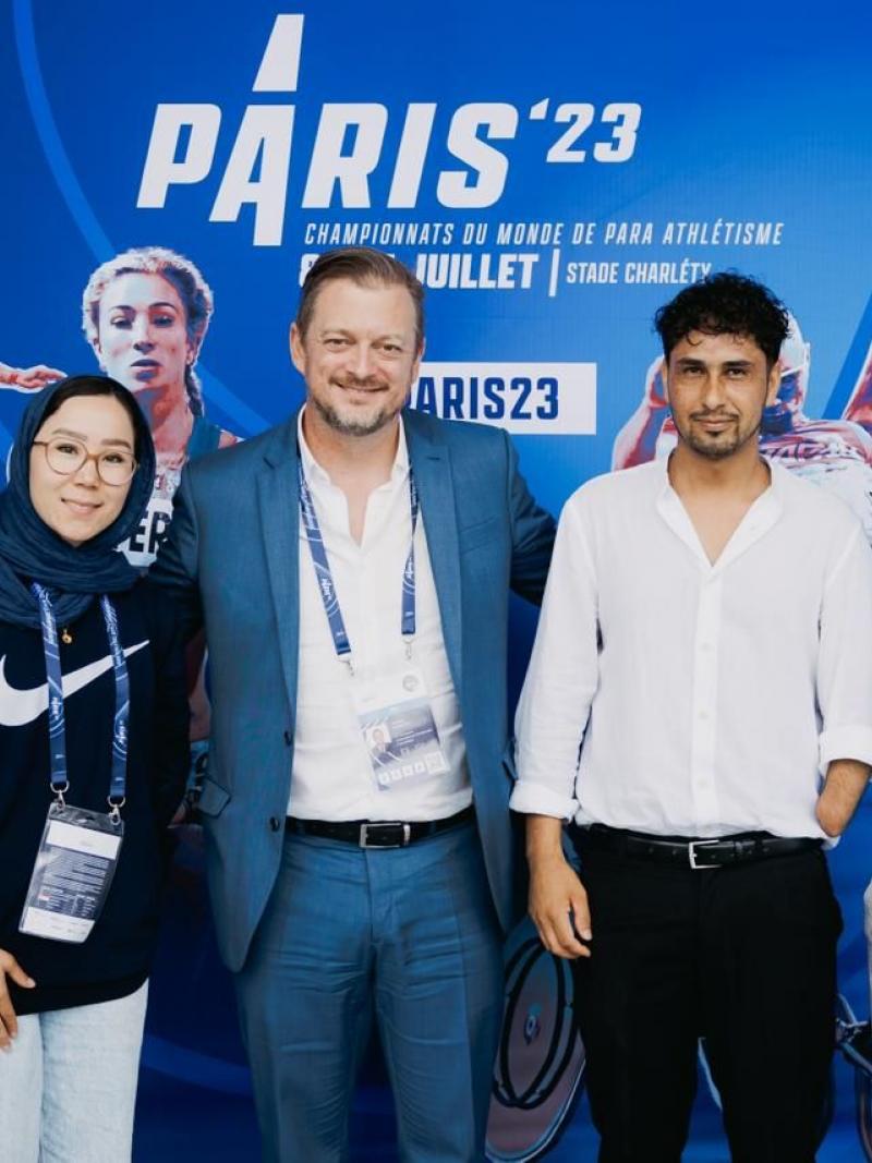 Five people pose for a group photograph in front of a Paris 23 Para Athletics World Championships banner