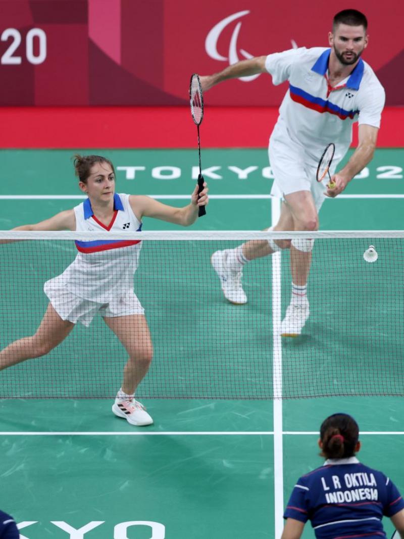 Photo shows a mixed doubles badminton match. A female athlete runs to a shuttle, in front of a male athlete.
