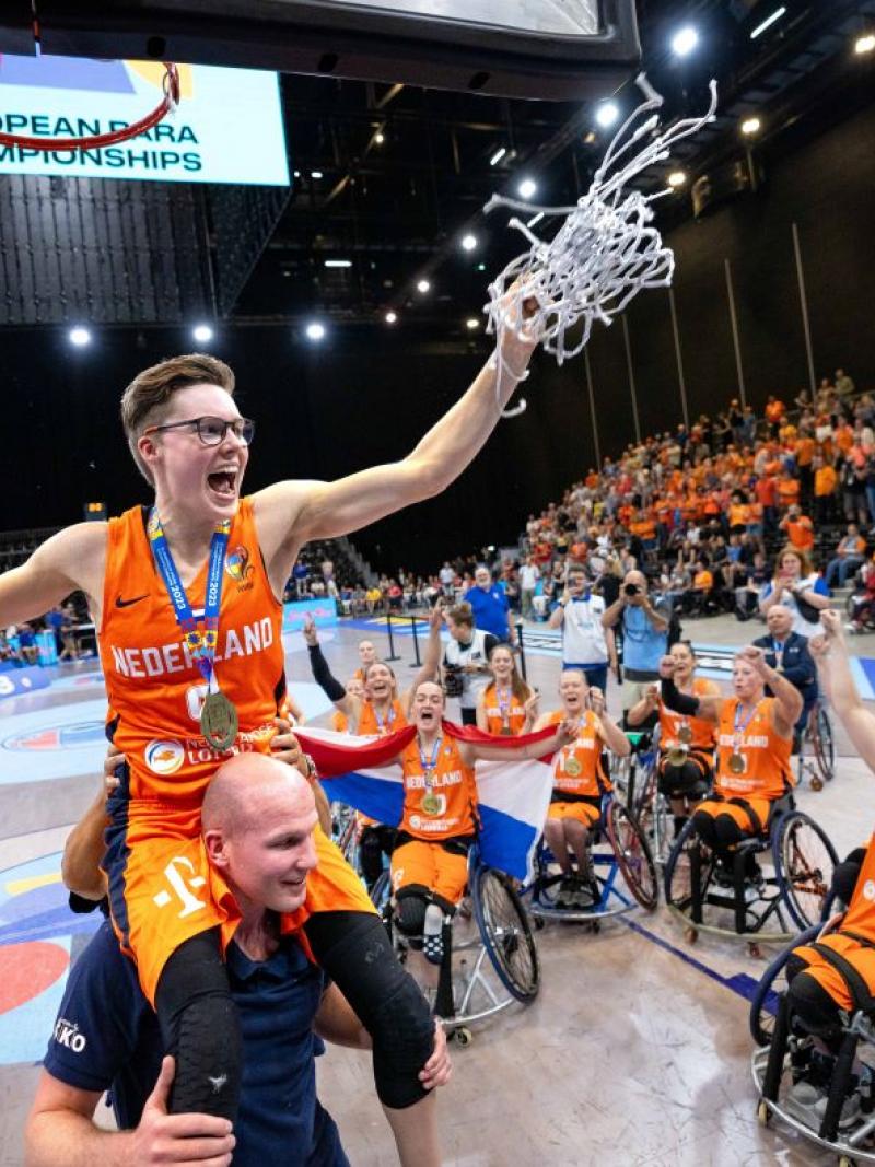 A female wheelchair basketball player cuts a basketball net, her teammates celebrate
