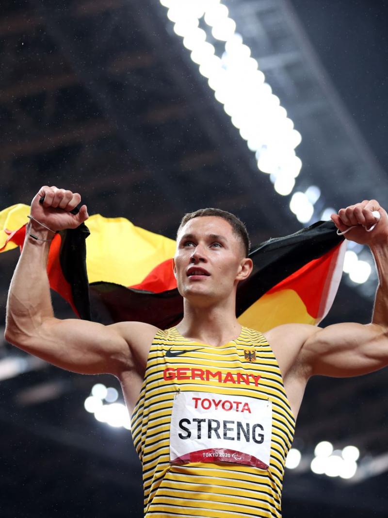 A male athlete holds the German flag with both hands at a stadium.
