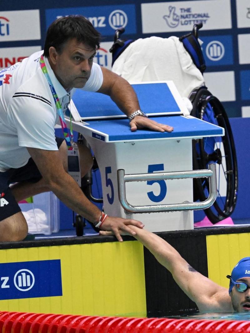 A male coach sits beside the pool and places his hand on a male swimmer's hand.