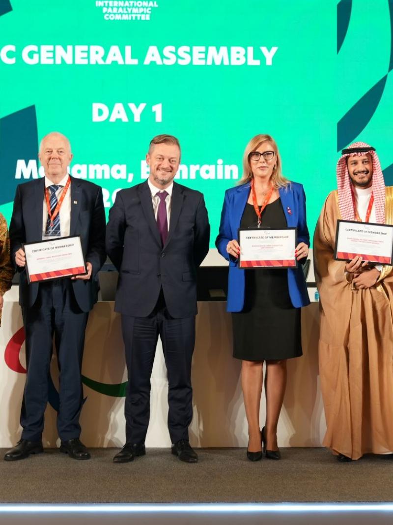 Five people pose for a group photo in front of a green screen that says "IPC General Assembly".