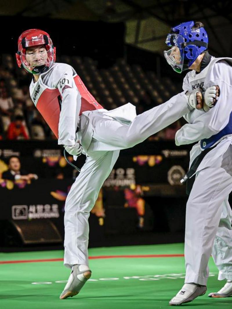 Two male Para taekwondo athletes fight in a match. An athlete wearing a red head gear kicks his opponent wearing a blue head gear.