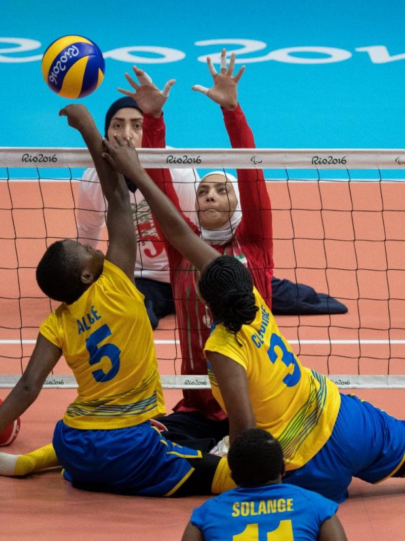 Two women's sitting volleyball teams playing at the Rio 2016 Paralympics. Two female players in yellow uniform are hitting the ball over the net, while two female players in red uniform are trying to block the ball.