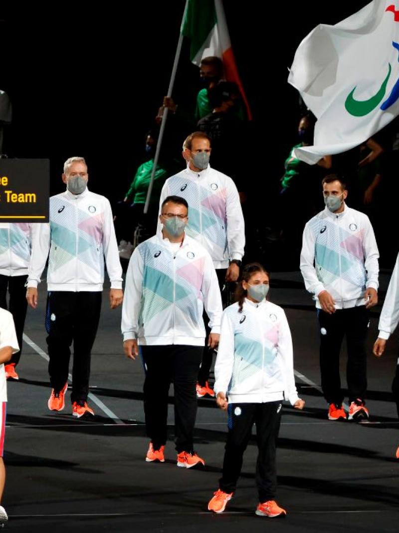 Ten athletes and officials take part in the athletes' parade at the opening ceremony of the Tokyo 2020 Paralympic Games. A volunteer is holding a sign that says "Refugee Paralympic Team" and a male athlete is carrying a flag of the Three Agitos.