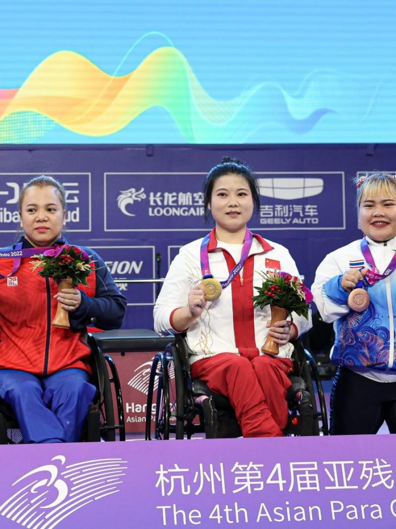 Two women in wheelchairs next to a short stature woman on a podium at the Asian Para Games