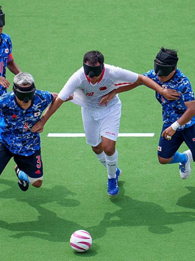 A blind football match between Japan and China at the Tokyo 2020 Paralympics. Three Japanese athletes wearing a blue jersey are trying to defend the ball from a Chinese athlete in a white jersey.