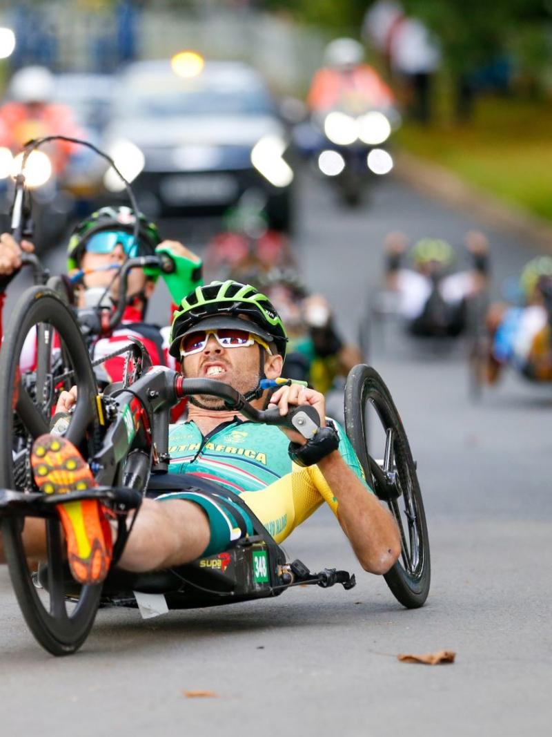 Six athletes compete on handcycles. The athlete leading the pack is wearing a South African jersey.