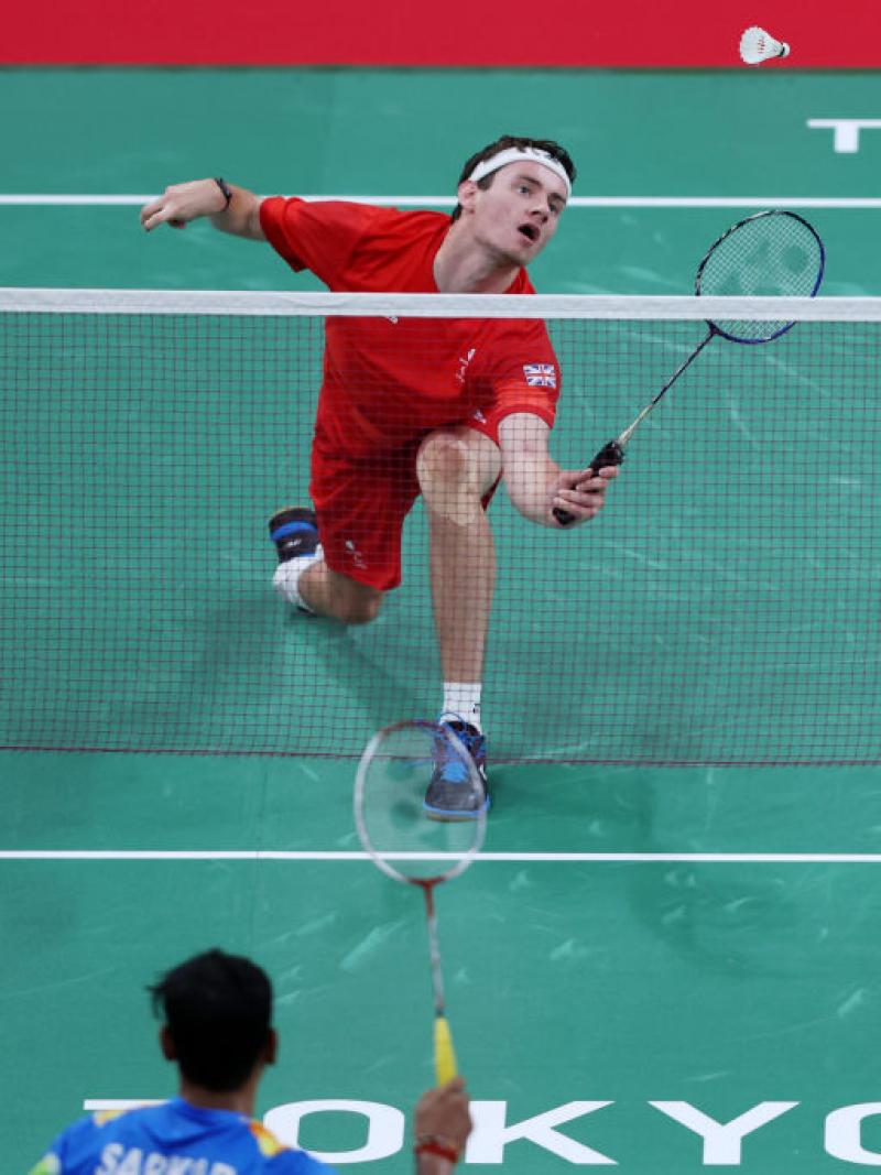 A Para badminton match between two male athletes at Tokyo 2020. A player in red uniform prepares to return the shuttle near the net.