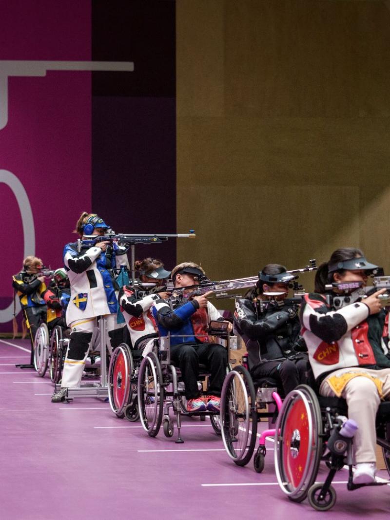 Six female athletes compete in Para shooting competition. Five athletes are competing in a wheelchair, while one athlete is standing.