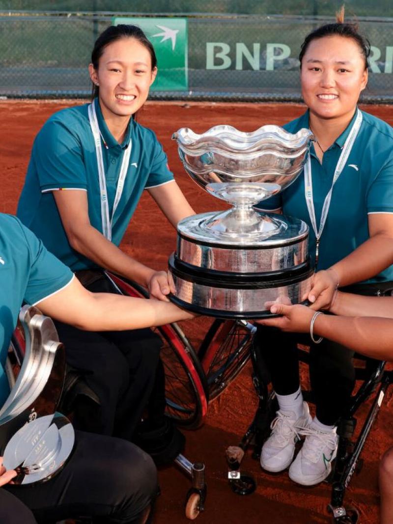 Four female wheelchair tennis athletes pose for a photograph. They are holding a silver trophy.