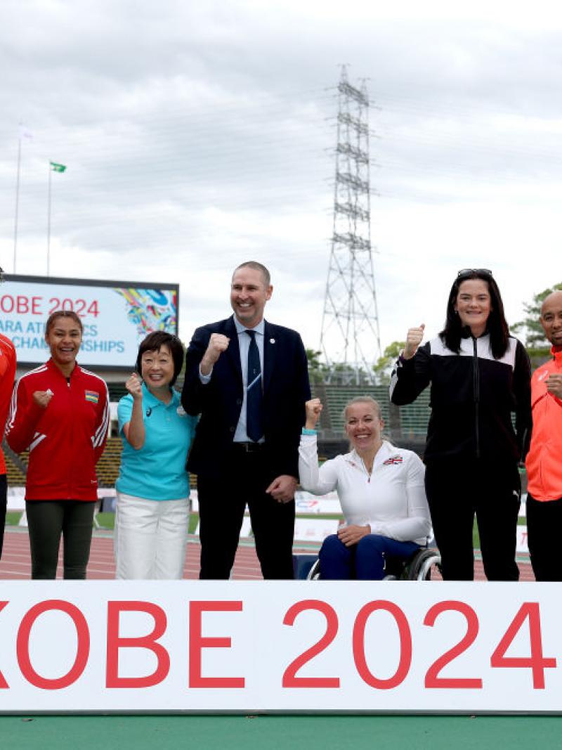 Seven standing person and a wheelchair user posing for a picture in an athletics track