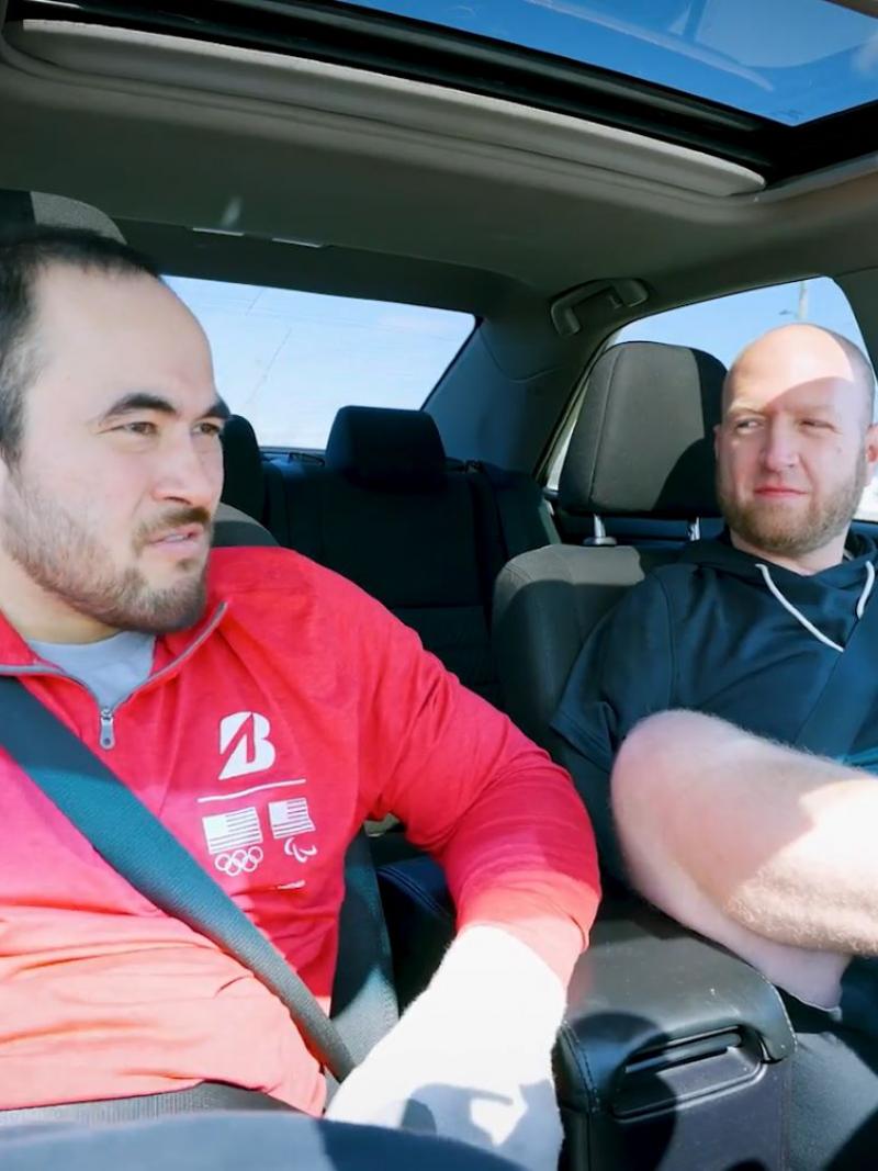 Two Para athletes are sitting in a car. One athlete is driving the car with his foot