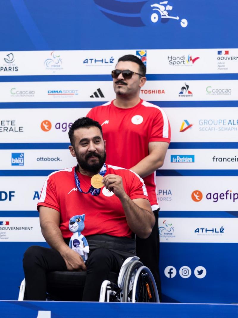 A man in a wheelchair showing a medal with a man standing behind him