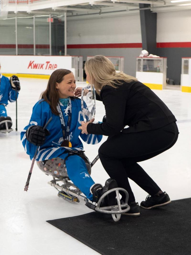 A female Para ice hockey player receiving a trophy from a woman in a hockey ring