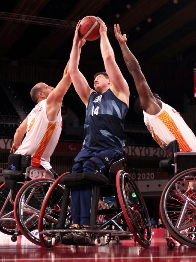 A male wheelchair basketball player wearing a blue jersey tries to shoot, while two players wearing a white jersey are reaching out to block him.
