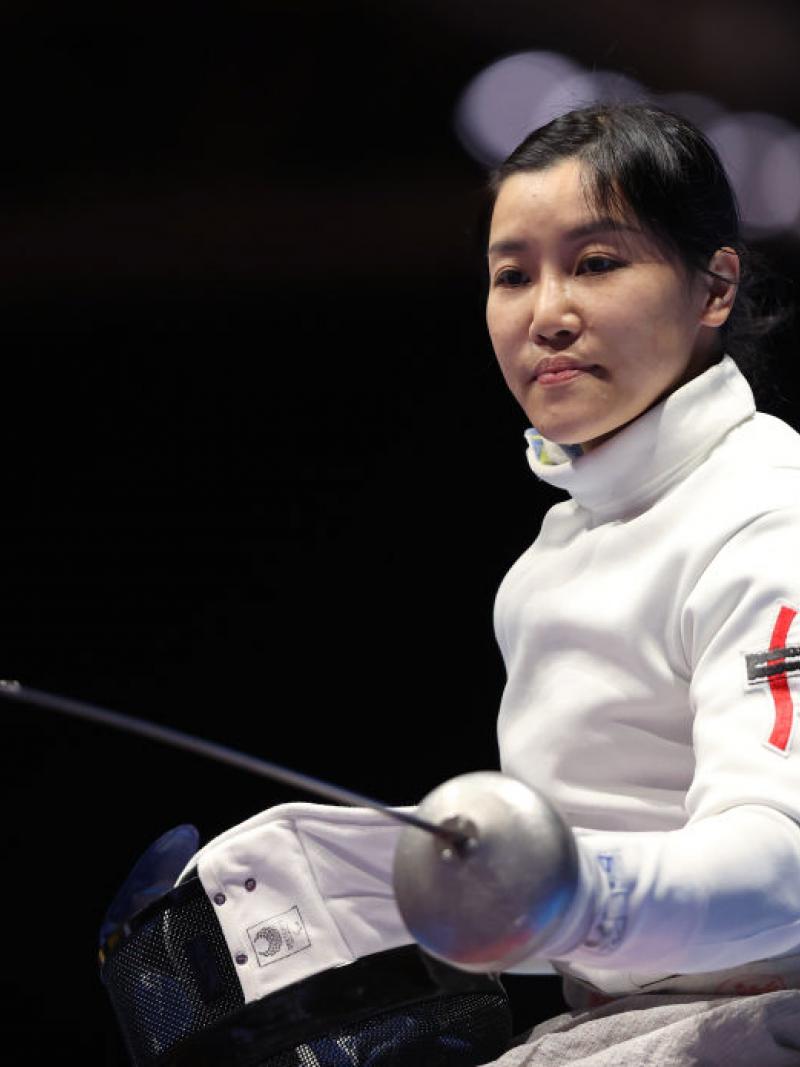 A female wheelchair fencer strikes a pose. She is holding her sword with her left and her mask with her right