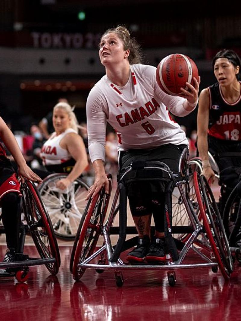 A female wheelchair basketball player carries the ball, while other players surround her