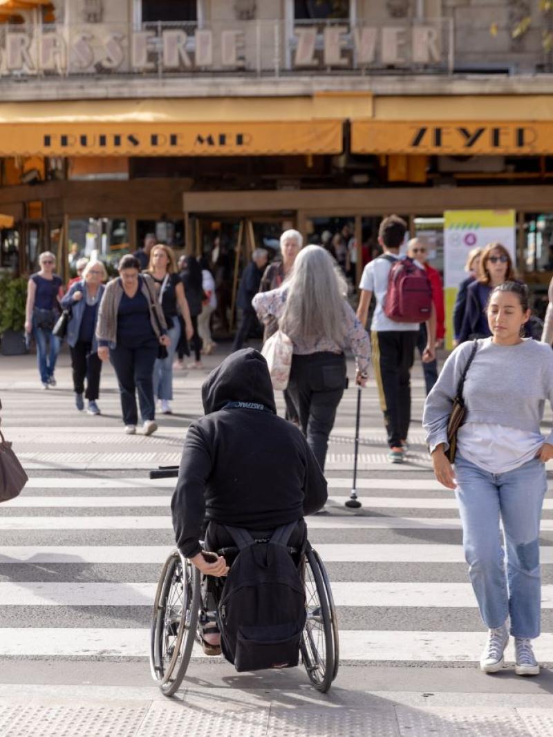 Many people cross a sidewalk in Paris. There are people in a wheelchair and people using canes.