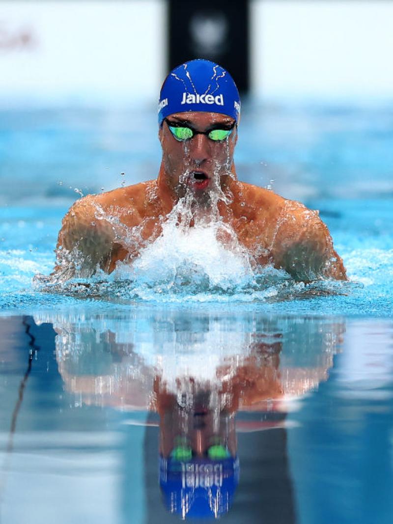 The head and shoulders of a swimmer in a blue cap and googles emerges from the water with a splash