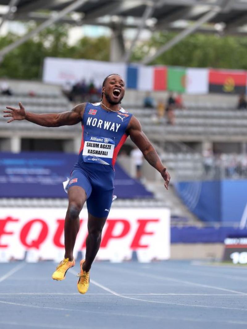 Three men cross the finish line on an athletics track. The man in the middle screams and has his arms outstretched.