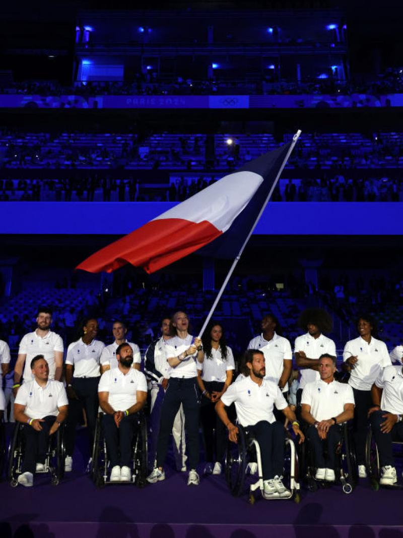 About 20 Paralympic athletes taking part in the Closing Ceremony of the Olympic Games. The female athlete in the centre is waving the French flag.