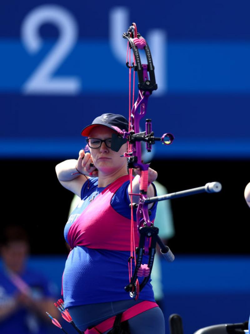 Jodie on the left in pink and blue with an obvious baby bump and Phoebe Paterson Pine on the right in blue Great Britain t-shirt both raise their bows to eye level to line up a shot