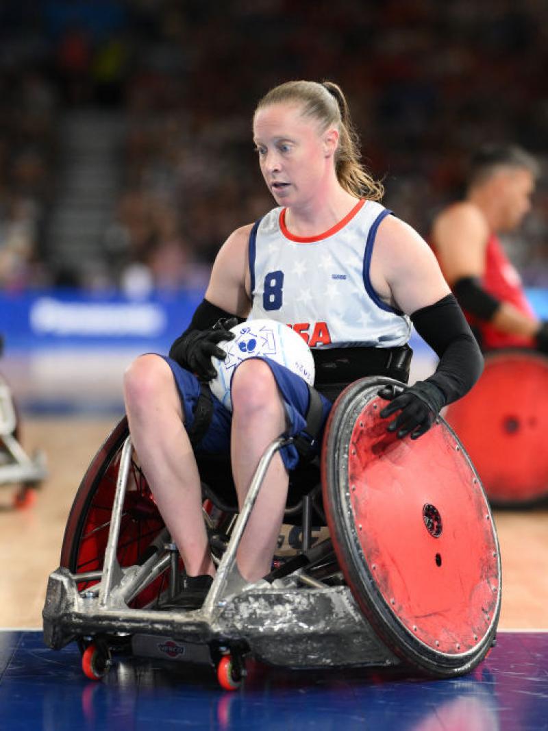 Woman in a white USA tea in wheelchair with red wheels holding a rugby ball