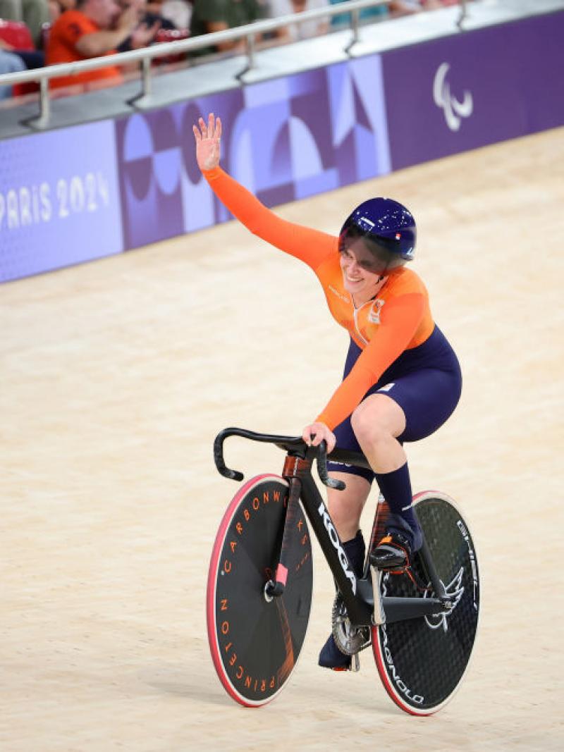 Caroline in orange with a blue helmet waves to crowd from her bike on the velodrome