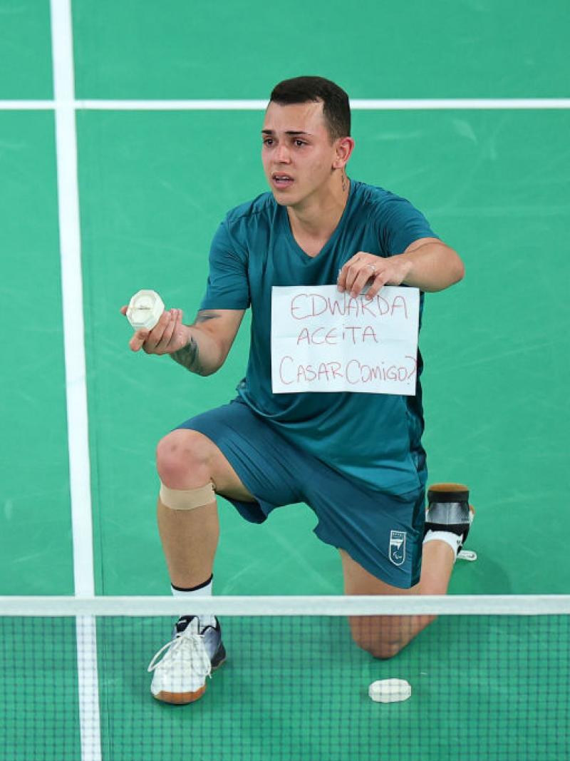 Man in blue tee and shorts kneels on the badminton court holding a ring