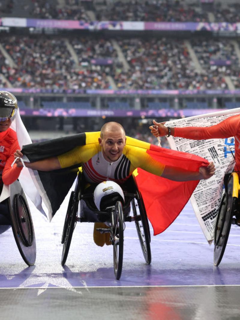 Three athletes in wheelchairs pose for the camera with the flags of their countries (Japan and Belgium) draped around their shoulders