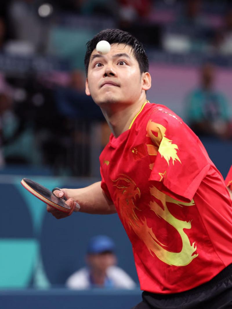 Two athletes play table tennis on the same team, a man looks at the ball and prepares to hit it, a woman braces herself to prepare for the next play