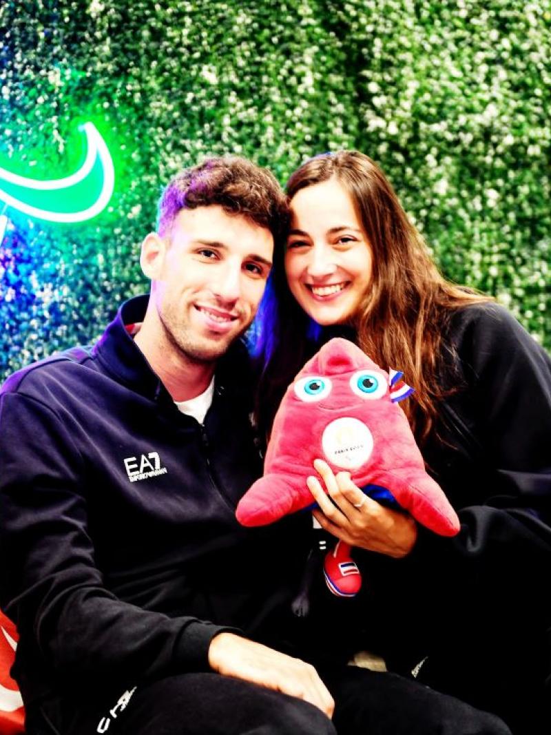 Man and woman face camera dressed in blue with Agitos in neon lights behind them holding the Paris 2024 mascot