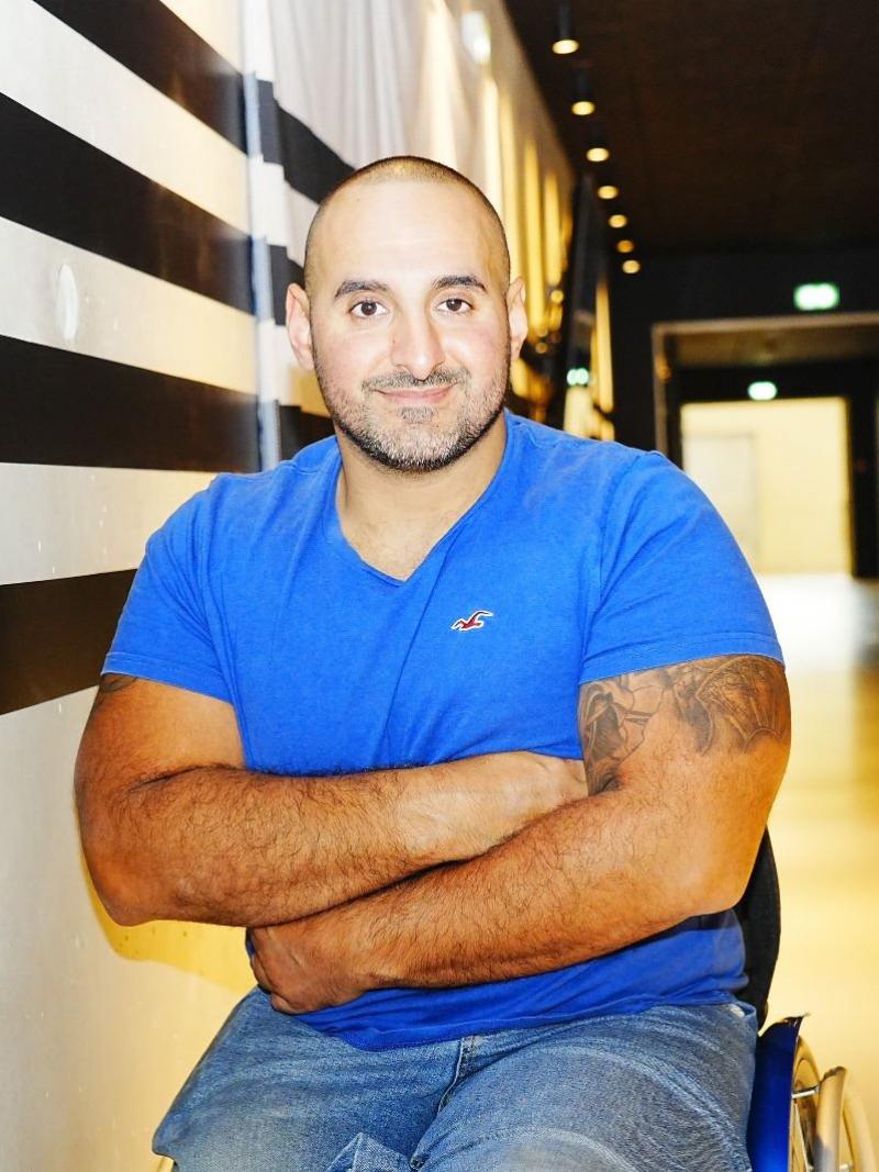 Man seated in blue t-shirt smiles to camera with arms folded