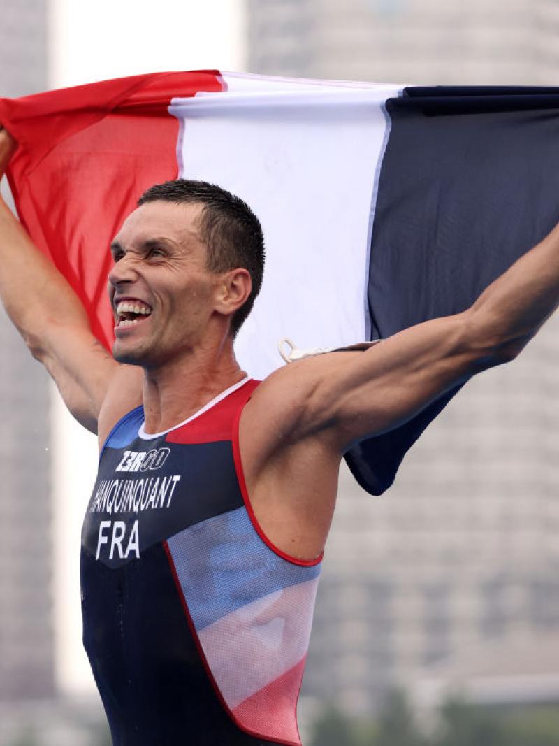 A man in a Team France triathlon uniform holds up the French flag and yells