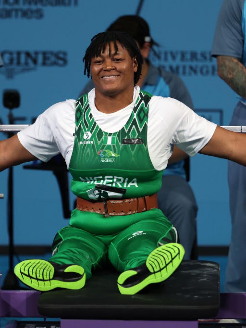 A woman in a green Team Nigeria uniform sits up on a Para powerlifting bench and stretches her arms out wide, smiling