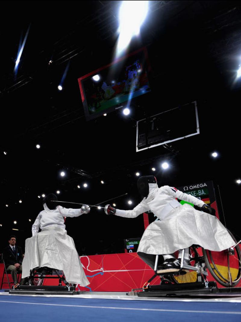 Two athletes in wheelchairs fence. They wear white uniforms and black face shields, their swords are crossed.