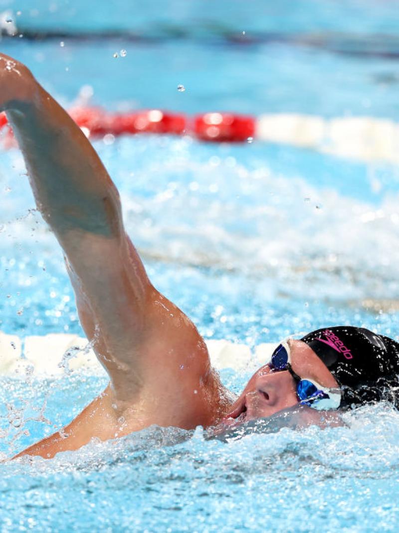 A man swimming reaches an arm out of the water and tilts his head up to take a breath