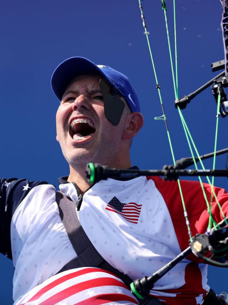 A man raises both arms in celebration, holding his archery bow in one hand