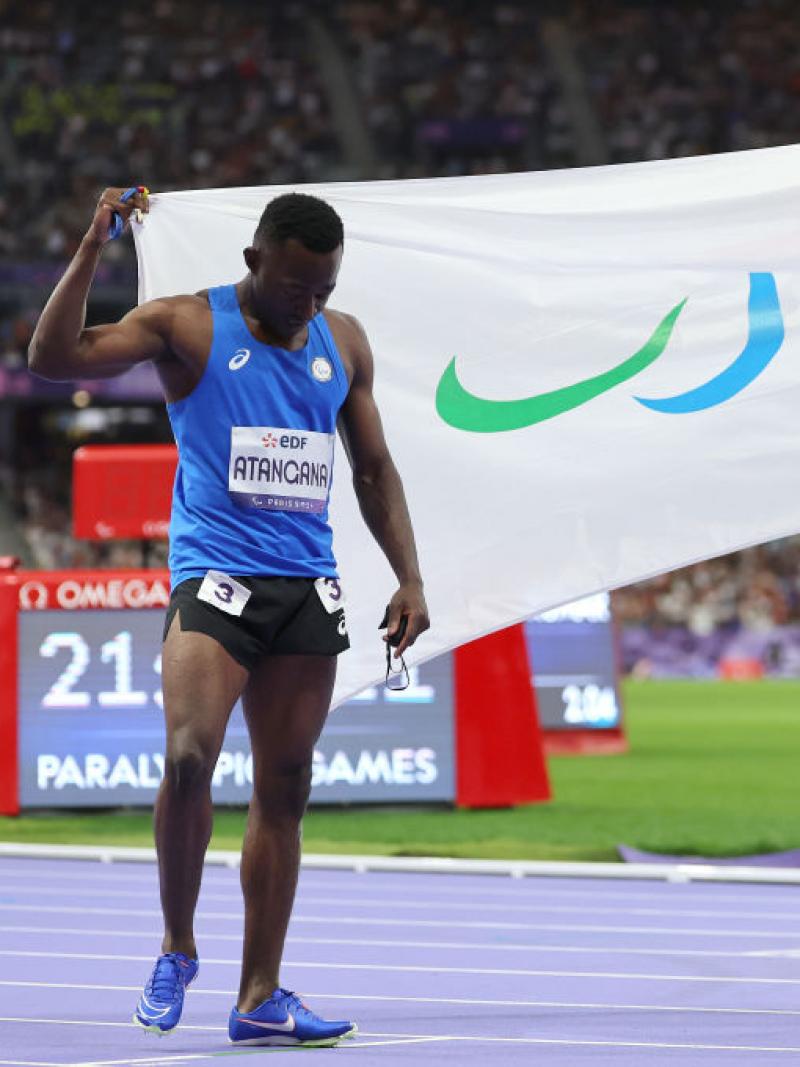 Two sprinters carry opposite sides of a white flag that has the Paralympic Agitos on it