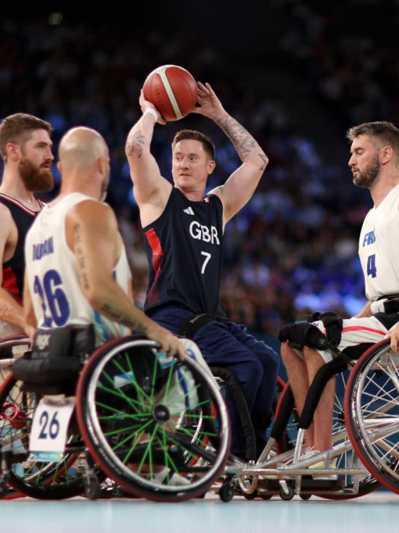 A man in a wheelchair holds a basketball above his head, surrounded by three other wheelchair basketball players
